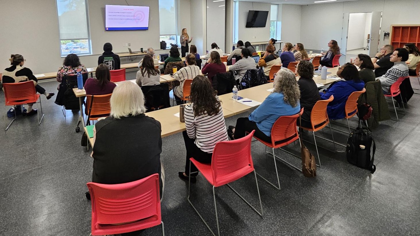 View of full room of participants seated and watching presenter in front of screen