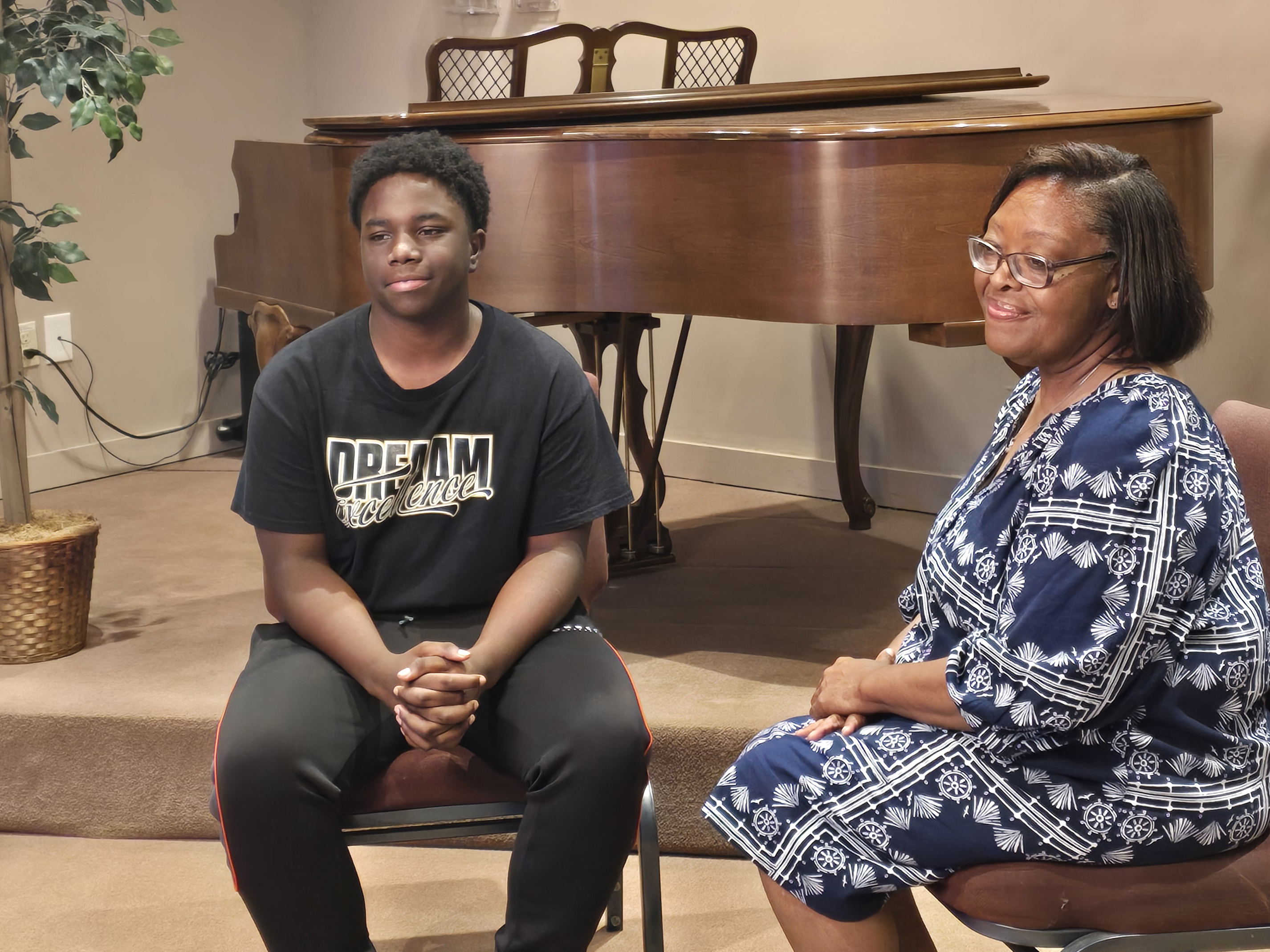 a teenage boy and older adult female sit in chairs with a grand piano in the background