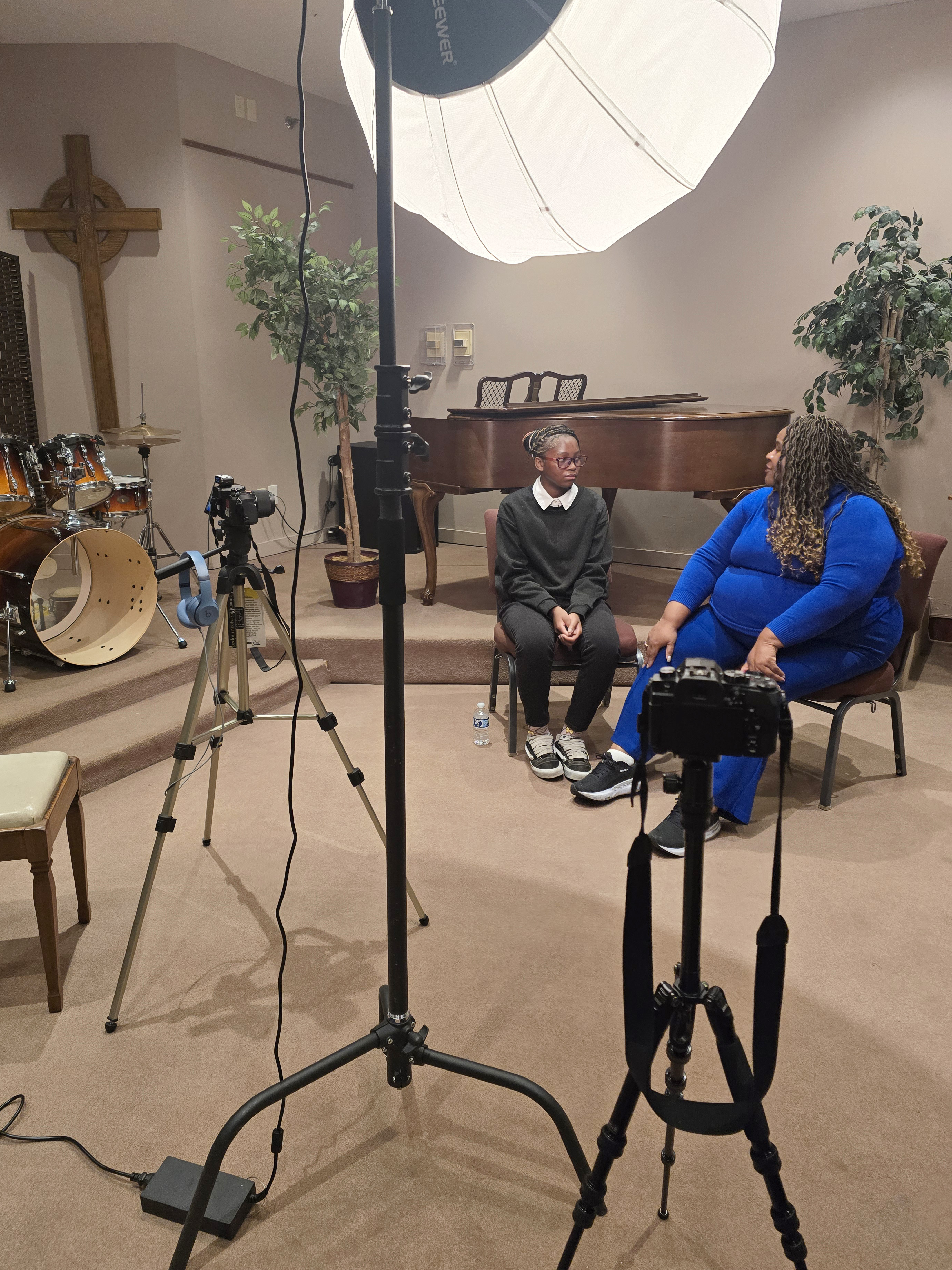 two cameras and an umbrella light, each on tripods in the foreground with a teenage girl and middle-age woman sitting in chairs in the background