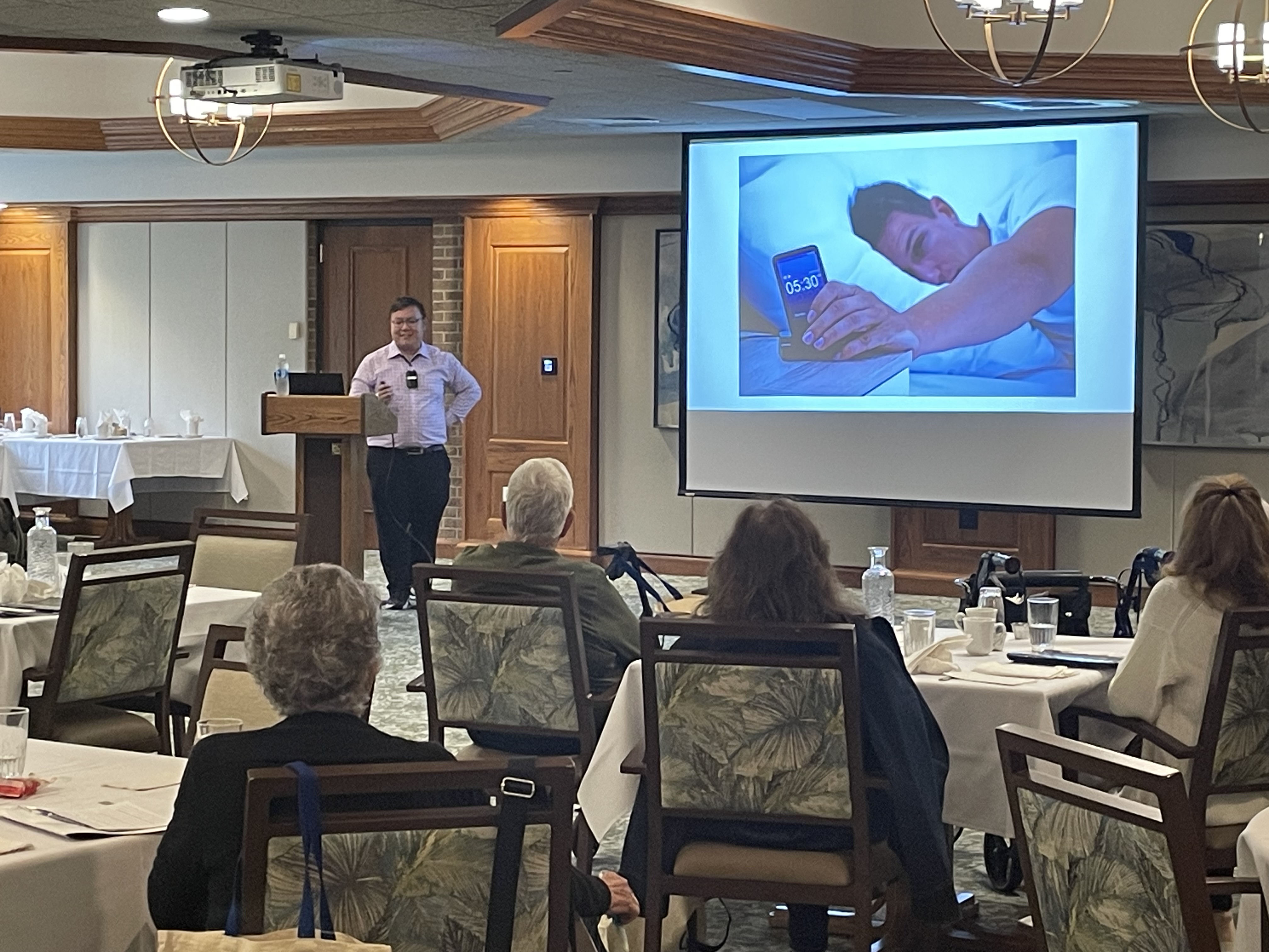 Presenter stands at podium beside large screen in a room full of older adults