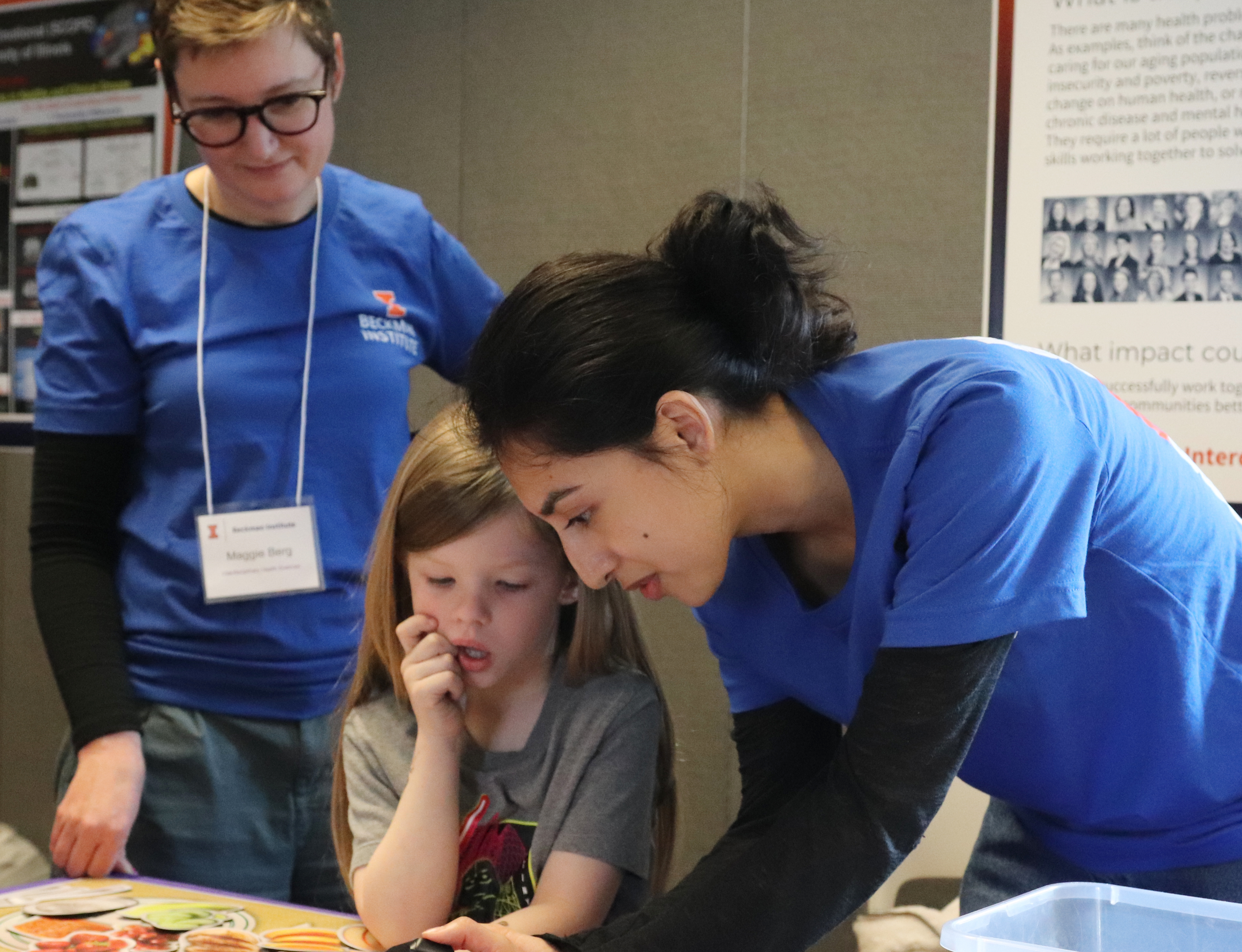 Child and student lean over a table next to a magnetic toy placement with foods while staff stands to the side observing.