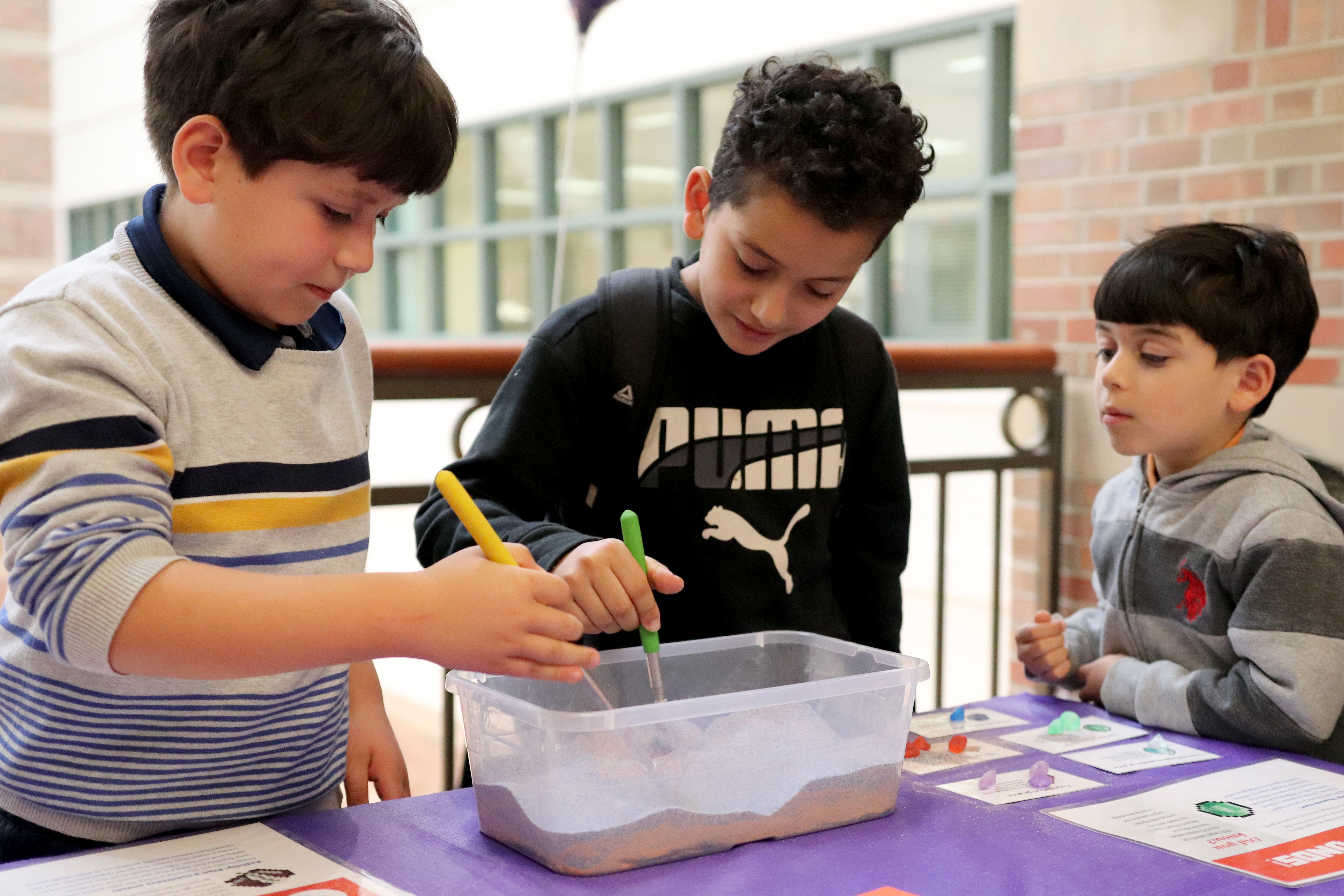 Two children use rakes to find gems in a clear bin of sand while younger child watches.