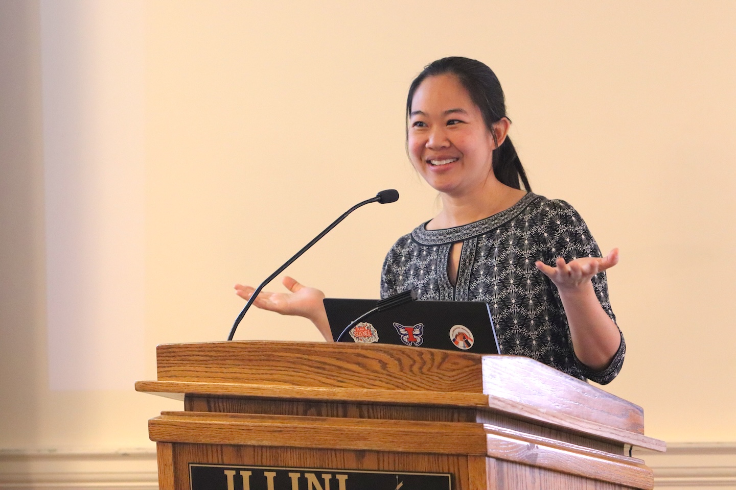 Pinshane Huang, presenting from a lectern, gestures and smiles at an unseen audience.