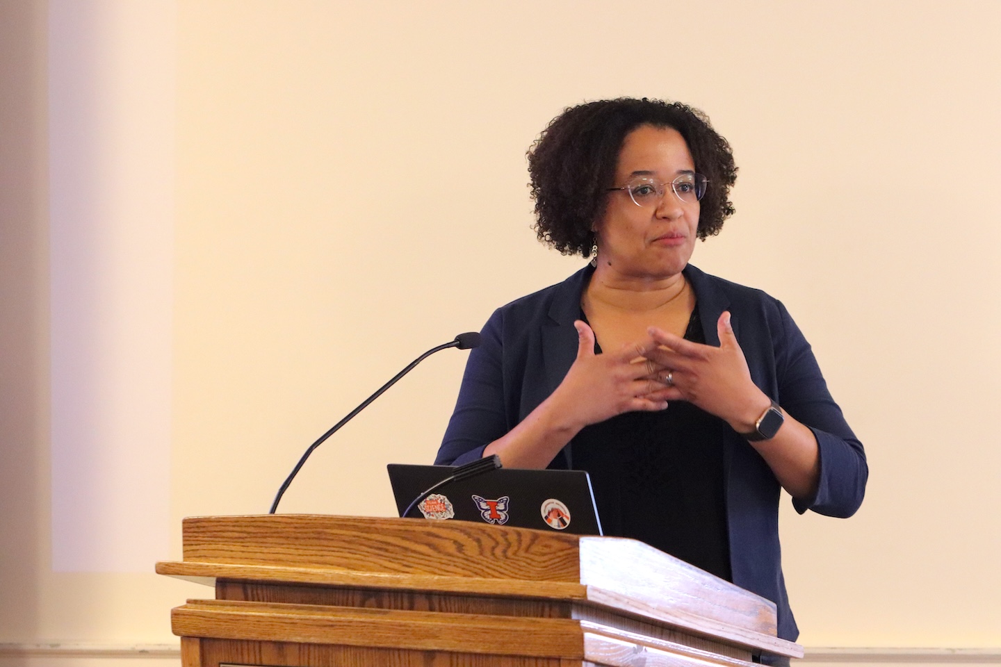 Deanna Hence, presenting from a lectern, gestures to herself while speaking to an unseen audience.